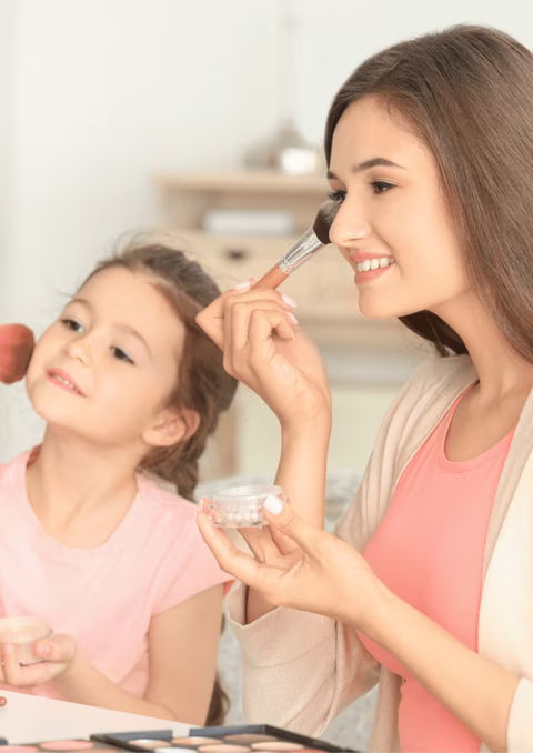 Une scène chaleureuse et joyeuse de complicité entre une mère et sa jeune fille pendant un moment de maquillage. La mère, souriante et concentrée, applique délicatement du maquillage sur le visage de la petite fille, qui semble ravie et excitée par l'attention. La mère tient un pinceau de maquillage et un petit pot de poudre ou de brillant, suggérant une touche légère et ludique plutôt qu'un maquillage complet. La fillette, avec un sourire doux et un regard attentif, semble apprécier ce moment de tendresse. Le décor autour d'elles est clair et domestique, probablement leur maison, ce qui donne une ambiance décontractée et intime.