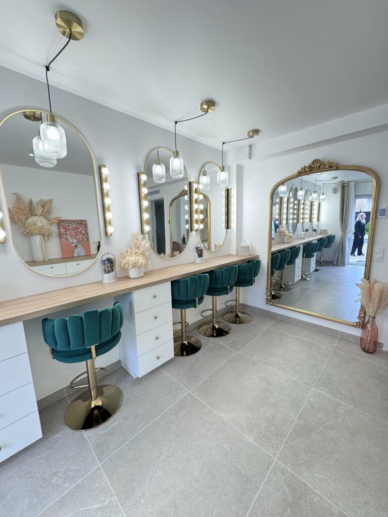 Vue intérieure du nouveau studio de maquillage de Jennifer Perseverante avec des stations de travail élégantes comprenant de grands miroirs ovales et des tabourets en velours vert émeraude. Une ambiance moderne et luxueuse caractérise l'espace, conçu pour offrir une expérience exceptionnelle de beauté