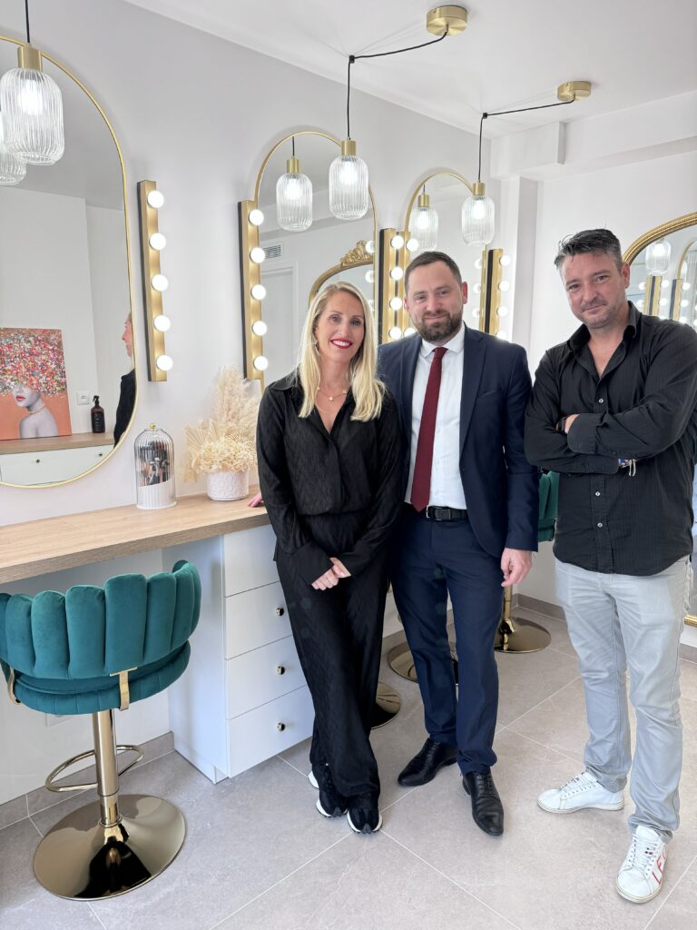 Jennifer Perseverante, accompagnée de Jonathan Wofsy, le Maire de Chevry-Cossigny, et d'Alexandre Chevalier, adjoint en charge de la vie économique, dans son élégant studio de maquillage. Le trio est entouré d'un décor sophistiqué, avec des miroirs illuminés et des tabourets en velours vert.