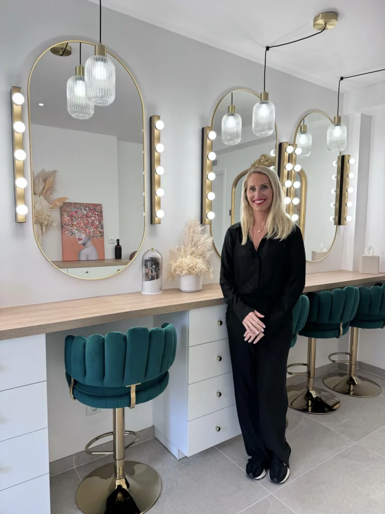 Jennifer Perseverante, maquilleuse professionnelle, souriante lors de l'inauguration de son studio de maquillage à Chevry-Cossigny. Elle se tient dans un espace élégant avec des miroirs illuminés et des tabourets en velours vert.