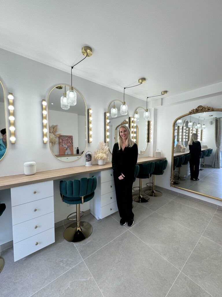 Jennifer Perseverante, maquilleuse professionnelle, souriante lors de l'inauguration de son studio de maquillage à Chevry-Cossigny. Elle se tient dans un espace élégant avec des miroirs illuminés et des tabourets en velours vert.