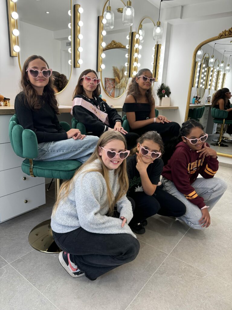 Un groupe de jeunes filles souriantes portant des lunettes en forme de cœur dans un élégant atelier de maquillage. Les miroirs illuminés et les fauteuils verts créent une ambiance chaleureuse et glamour, idéale pour un moment de partage et de créativité