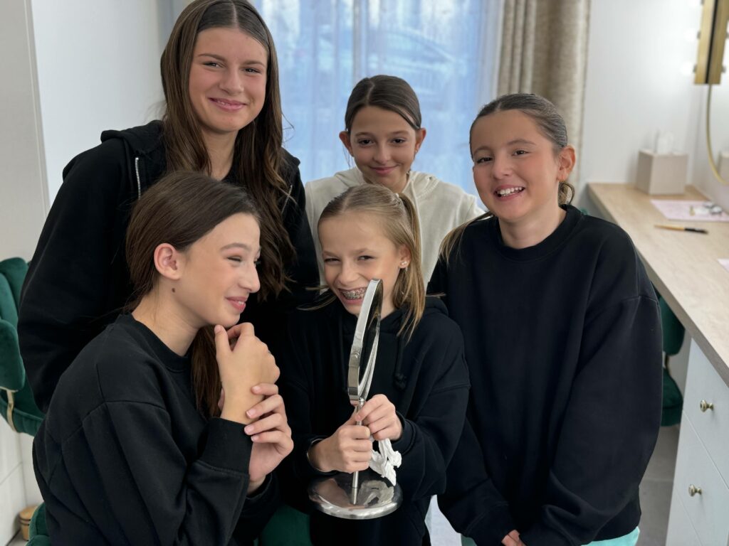 Cinq jeunes filles souriantes posant ensemble dans un atelier de maquillage moderne. L'une d'elles tient un miroir élégant, reflétant une ambiance conviviale et joyeuse dans un espace chaleureux et bien aménagé.